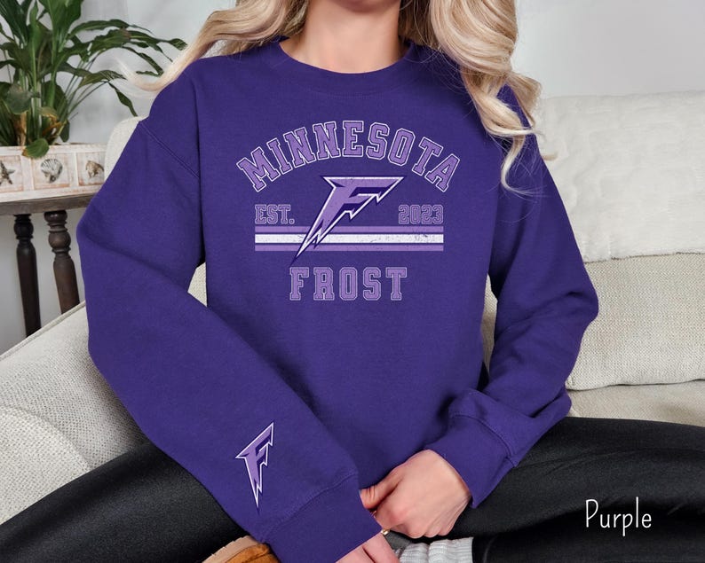 Pode incluir: Camisola roxa com um design gr&aacute;fico branco e roxo. O gr&aacute;fico apresenta um raio com o texto "Minnesota Frost Est. 2023".