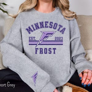 Pode incluir: Uma camisa de moletom cinza com um design gr&aacute;fico roxo que diz "Minnesota Frost" com o texto "Est. 2023" embaixo.