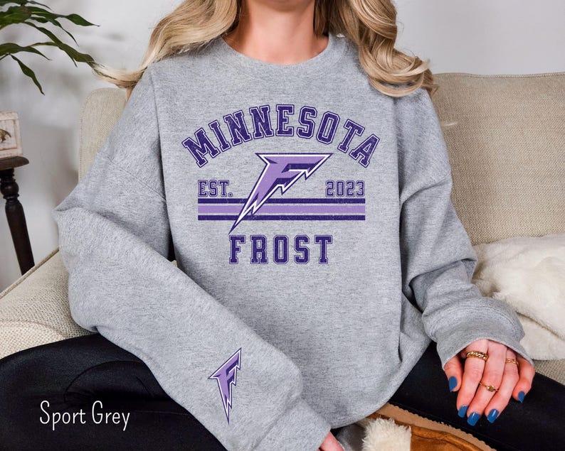 Pode incluir: Uma camisa de moletom cinza com um design gr&aacute;fico roxo que diz "Minnesota Frost" com o texto "Est. 2023" embaixo.