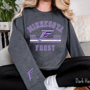 Pode incluir: Uma camisa de manga comprida cinza escuro com um gr&aacute;fico roxo de um raio e o texto "Minnesota Frost Est. 2003".