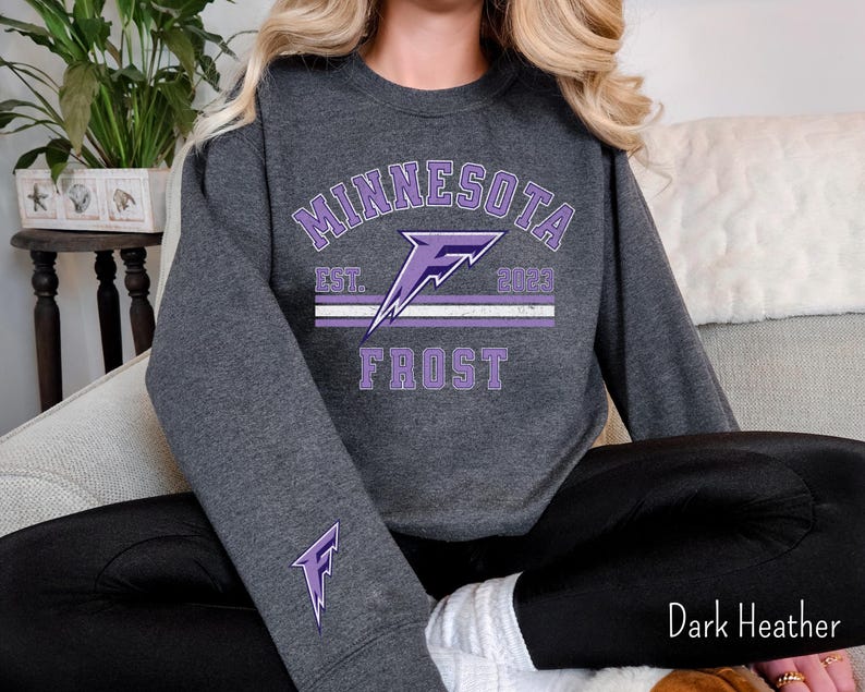 Pode incluir: Uma camisa de manga comprida cinza escuro com um gr&aacute;fico roxo de um raio e o texto "Minnesota Frost Est. 2003".