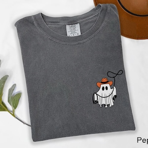 K&ouml;nnte beinhalten: Dunkelgraues T-Shirt mit Rundhalsausschnitt und kurzen &Auml;rmeln. Das Shirt zeigt eine Grafik eines Geistes mit Cowboyhut und Lasso. Ein brauner Cowboyhut ist in der oberen rechten Ecke sichtbar.