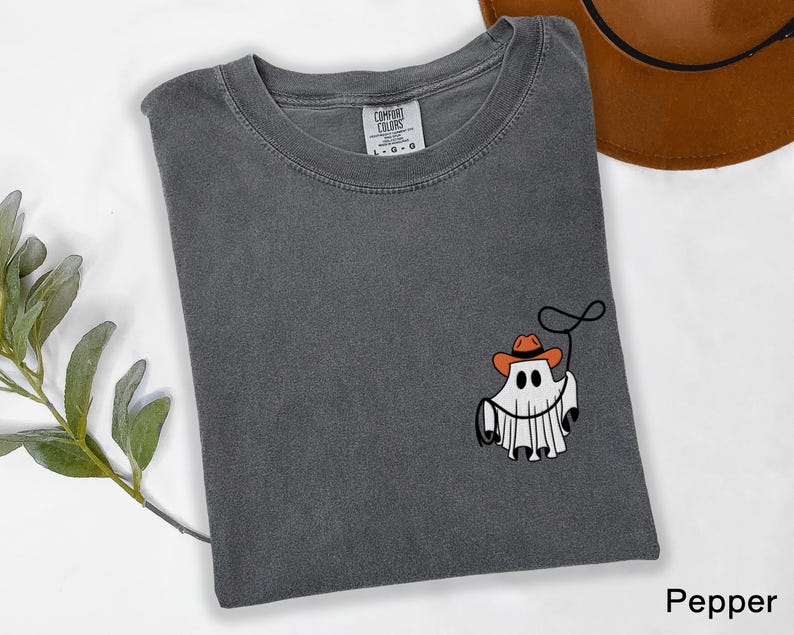 K&ouml;nnte beinhalten: Dunkelgraues T-Shirt mit Rundhalsausschnitt und kurzen &Auml;rmeln. Das Shirt zeigt eine Grafik eines Geistes mit Cowboyhut und Lasso. Ein brauner Cowboyhut ist in der oberen rechten Ecke sichtbar.