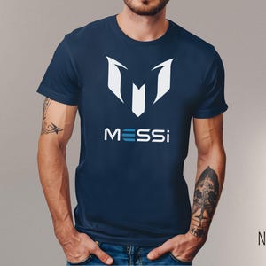 May include: Navy blue t-shirt with a white graphic of a stylized "M" and the word "MESSI" in blue and white letters.