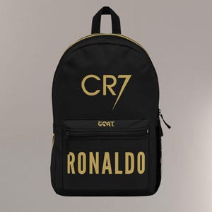 May include: Black backpack with gold lettering. The text includes "CR7", "GOAT", and "RONALDO". The backpack has a top handle, a front pocket, and a side mesh pocket. The zipper is black.