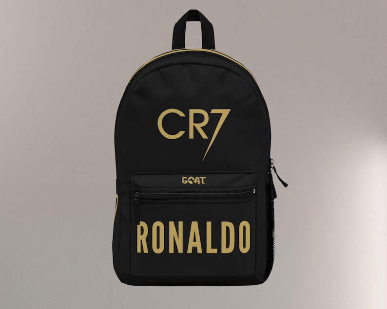 May include: Black backpack with gold lettering. The text includes "CR7", "GOAT", and "RONALDO". The backpack has a top handle, a front pocket, and a side mesh pocket. The zipper is black.
