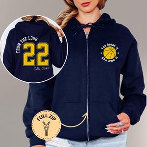 May include: Navy blue full-zip hoodie with a basketball graphic and the text "You break it, you own it." The back of the hoodie features the number 22 in yellow with the text "From the logo" and the name "Caitlin Clark22".