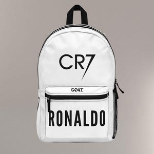 May include: White backpack with black accents, featuring the text "CR7" and "RONALDO" in bold black letters. The word "GOAT" is also printed on the front pocket. Includes a black handle and a side mesh pocket.