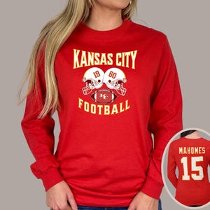 May include: Red long-sleeve t-shirt with the text "Kansas City Football" and a graphic of two football helmets with a football in the middle. The helmets have the numbers 19 and 60 on them.