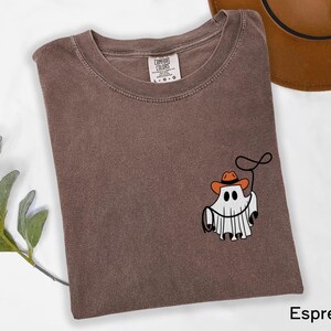 K&ouml;nnte beinhalten: Espressofarbenes T-Shirt mit einer Grafik eines Geistes, der einen Cowboyhut tr&auml;gt und ein Lasso h&auml;lt. Das Shirt besteht aus weichem Material und hat einen Rundhalsausschnitt. Das Comfort Colors-Etikett ist innen am Kragen sichtbar.