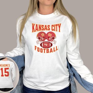 May include: White long-sleeve shirt with the text "Kansas City Football" and a graphic of two football helmets with the numbers 19 and 60. The helmets are red with gold accents and are facing each other. A football with the letters "KC" is in the middle of the helmets.