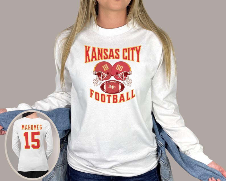 May include: White long-sleeve shirt with the text "Kansas City Football" and a graphic of two football helmets with the numbers 19 and 60. The helmets are red with gold accents and are facing each other. A football with the letters "KC" is in the middle of the helmets.