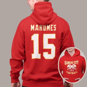May include: Red hooded sweatshirt with the name "Mahomes" and the number "15" printed on the back in white. The front of the sweatshirt has a smaller image of two football helmets with the text "Kansas City Football" printed below.