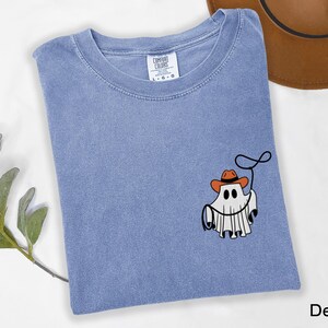 K&ouml;nnte beinhalten: Hellblaues Denim-T-Shirt mit einem Cartoon-Geist, der einen Cowboyhut tr&auml;gt und ein Lasso h&auml;lt. Das Shirt hat einen Rundhalsausschnitt und kurze &Auml;rmel. Das Wort "Denim" ist unten rechts aufgedruckt.