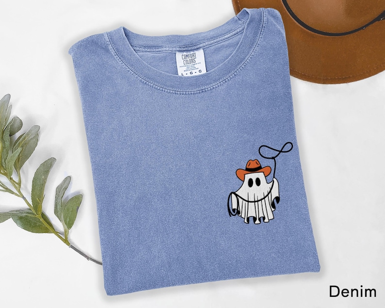 K&ouml;nnte beinhalten: Hellblaues Denim-T-Shirt mit einem Cartoon-Geist, der einen Cowboyhut tr&auml;gt und ein Lasso h&auml;lt. Das Shirt hat einen Rundhalsausschnitt und kurze &Auml;rmel. Das Wort "Denim" ist unten rechts aufgedruckt.