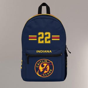 May include: Navy blue backpack with a black handle and zipper. The backpack features the number "22" in yellow with red stripes, and the word "INDIANA" in yellow. A circular logo with the text "CAITLIN CLARK" is also visible.