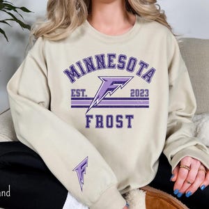 Pode incluir: Uma camisa de moletom bege com um gr&aacute;fico roxo de um raio e o texto "Minnesota Frost Est. 2023".