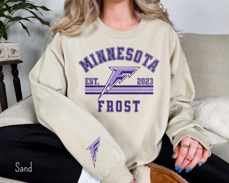 Pode incluir: Uma camisa de moletom bege com um gr&aacute;fico roxo de um raio e o texto "Minnesota Frost Est. 2023".