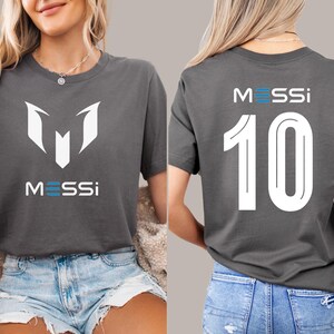 May include: A gray t-shirt with a white graphic of the letter "M" and the word "MESSI" on the front. The back of the shirt has the number "10" and the word "MESSI" in white.