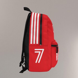 May include: Red backpack with white stripes and the number 7 in white. The backpack has a black back and straps, a top handle, and a front pocket with a black zipper. The word "R" is visible on the side.