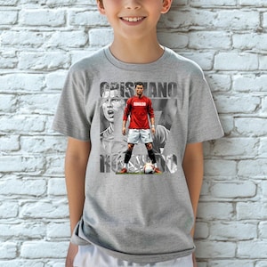 May include: A heather gray short-sleeve t-shirt featuring a graphic print of Cristiano Ronaldo in a red jersey and white shorts. The design includes a large black and white portrait background and the text CRISTIANO RONALDO in bold letters.