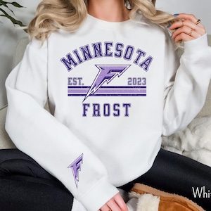 Pode incluir: Camisola branca com um design gr&aacute;fico roxo e branco que diz "Minnesota Frost" com um logotipo de raio e o texto "Est. 2023".