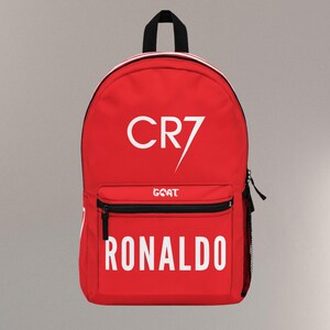May include: A red backpack with the text "CR7" and "RONALDO" in white. The backpack has a black zipper, a small front pocket, and a black handle. The word "GOAT" is printed below the "CR7" text.