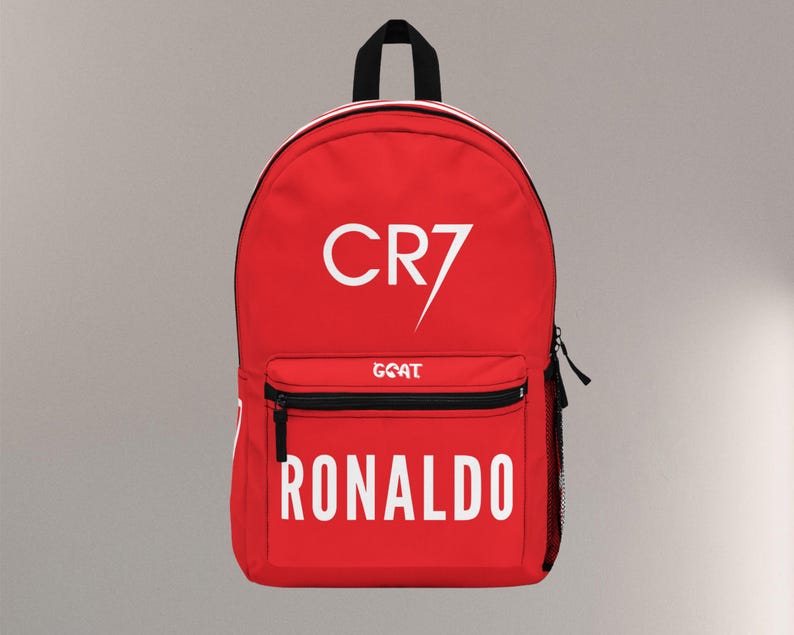 May include: A red backpack with the text "CR7" and "RONALDO" in white. The backpack has a black zipper, a small front pocket, and a black handle. The word "GOAT" is printed below the "CR7" text.