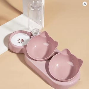 May include: A pink cat-shaped pet food and water bowl set. The set includes two bowls, a water dispenser, and a water reservoir with a paw print design. The bowls and base are a soft pink color, and the water bottle is clear.