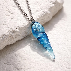 May include: A blue, spiral-shaped pendant necklace. The pendant is made of translucent glass and hangs from a silver-coloured chain. The necklace is displayed on a white, textured surface.
