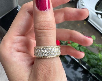 May include: A wide gold band ring featuring three horizontal rows of small, round, clear stones set closely together. The ring has a polished gold finish and covers a significant portion of the finger, showcasing a dense, sparkling pavé design.