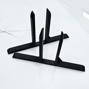 May include: Two black metal display stands. Each stand has a rectangular base with four vertical, pointed supports. The stands are designed to hold items and are set against a white marble-like surface.