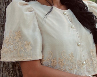 May include: A woman wearing a cream-colored blouse with delicate floral embroidery and pearl buttons. She is wearing a brown skirt and a pearl bracelet.