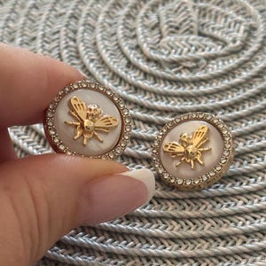 Puede incluir: Un par de pendientes de tono dorado con un diseño de abeja de nácar blanco y detalles de cristal transparente.