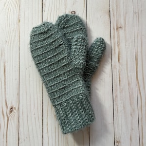 Peut inclure: Une paire de mitaines tricotées de couleur vert d'eau. Les mitaines présentent un motif côtelé texturé sur le dessus et un tricot plus doux sur les paumes et les poignets. Les mitaines sont présentées sur une surface en bois clair.