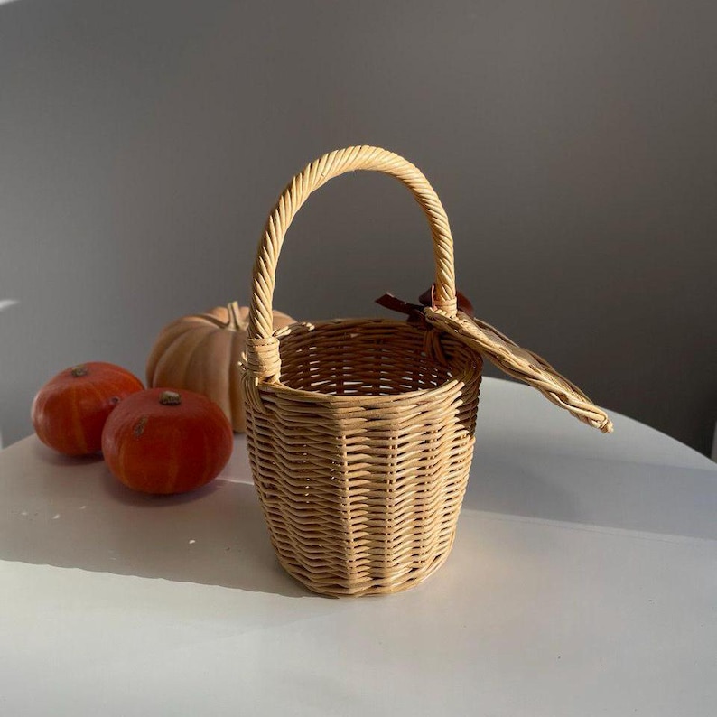 May include: A light brown wicker basket with a curved handle and a hinged lid. Two small orange pumpkins and one tan pumpkin are next to the basket. The basket sits on a white surface.