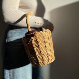 May include: A cylindrical wicker basket bag with a twisted handle and a brown leather detail. The bag is made of woven light brown wicker. The bag is held by a person wearing a white long-sleeve top and blue jeans.