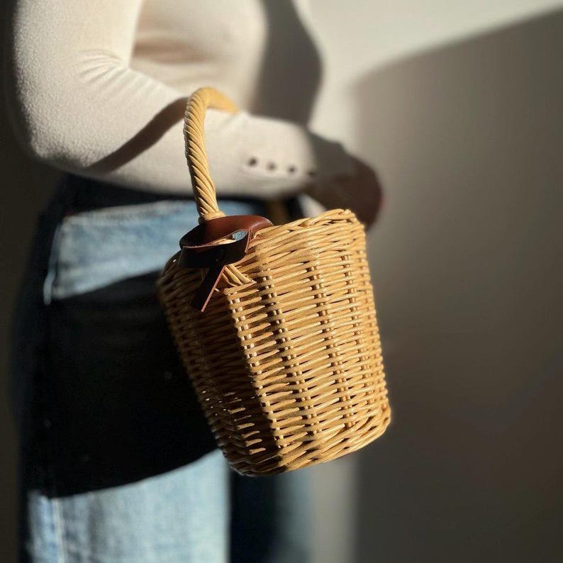 May include: A cylindrical wicker basket bag with a twisted handle and a brown leather detail. The bag is made of woven light brown wicker. The bag is held by a person wearing a white long-sleeve top and blue jeans.