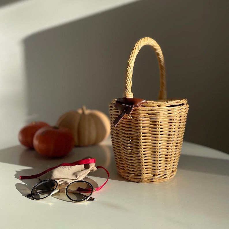 May include: A woven wicker basket with a curved handle, a brown leather accent, and a red drawstring bag with sunglasses. Two red apples and a small pumpkin are in the background.
