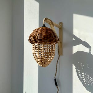 May include: A wicker acorn-shaped lampshade hangs from a wooden wall mount. The lampshade is made of natural woven wicker and has a warm, rustic aesthetic.
