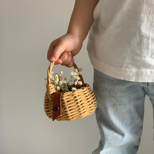 May include: A small woven basket with a brown leather handle and a button closure. The basket is filled with white flowers and is being held by a person.