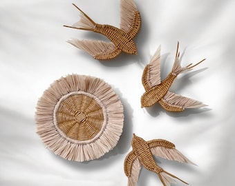 May include: Three woven wicker bird wall hangings with fringed edges in a light brown color. The birds are in a flying position and are arranged around a round woven wicker coaster with a fringed edge in a light pink color.