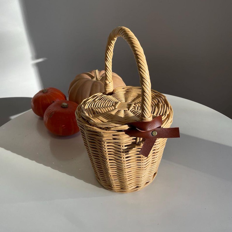 May include: A light brown wicker basket with a handle and a lid, fastened with a brown leather strap and bow. The basket is on a white surface, with three pumpkins in the background.