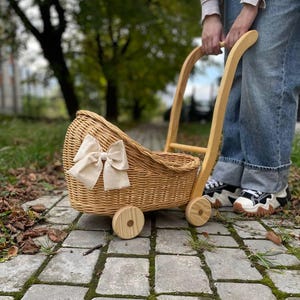 May include: A wooden doll's pram with a wicker basket and a white bow. The pram has two wheels and a handle. The pram is on a brick path.