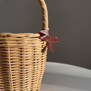 May include: A light brown woven wicker basket with a curved handle. A brown leather bow is tied near the handle. The basket's weave is tightly constructed, suggesting durability and a rustic aesthetic. The basket is suitable for picnics.