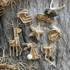 Könnte beinhalten: Eine Auswahl an Weihnachtsbaumschmuck aus geflochtenem Stroh, darunter ein Rentier, ein Stern, ein Herz, ein Schaukelpferd, ein Auto, ein Fäustling und ein Hase. Jedes Ornament ist mit einer Schleife und einer Schnur zum Aufhängen versehen. Grauer Hintergrund.