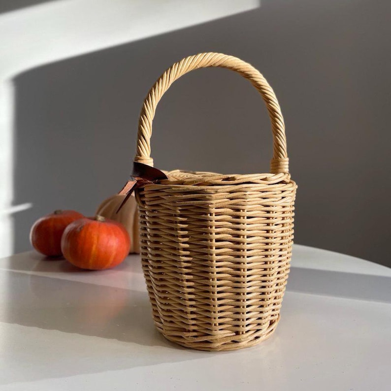 May include: A light brown wicker basket with a curved handle and a brown ribbon. Two orange pumpkins and a white gourd are in the background. The basket is cylindrical and appears to be empty.