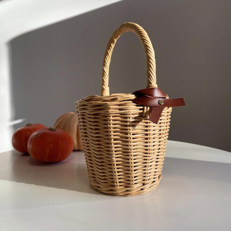 May include: A woven wicker basket with a curved handle and a brown leather accent. The basket is cylindrical and has a matching handle. Three small pumpkins are in the background.