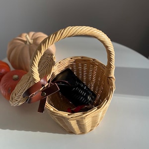 May include: A small, round, woven wicker basket with a handle. The basket contains a black wallet and a red item. A brown leather strap with a buckle secures the lid. A pumpkin and two small orange pumpkins are in the background.