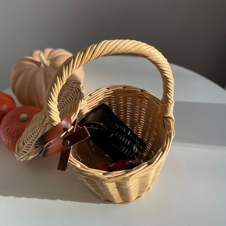 May include: A small, round, woven wicker basket with a handle. The basket contains a black wallet and a red item. A brown leather strap with a buckle secures the lid. A pumpkin and two small orange pumpkins are in the background.